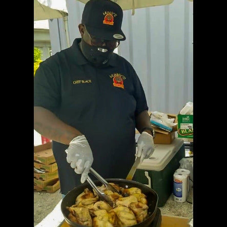 Person cooking food in a pan outdoors, wearing a black shirt and hat.